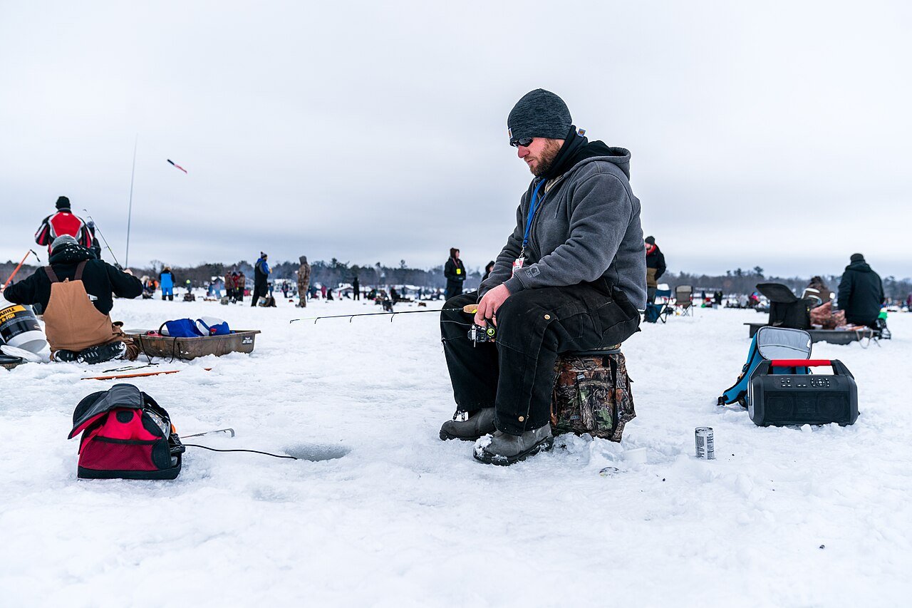 Angler sitting on a bucket ice fishing next to drilled holes on a frozen lake