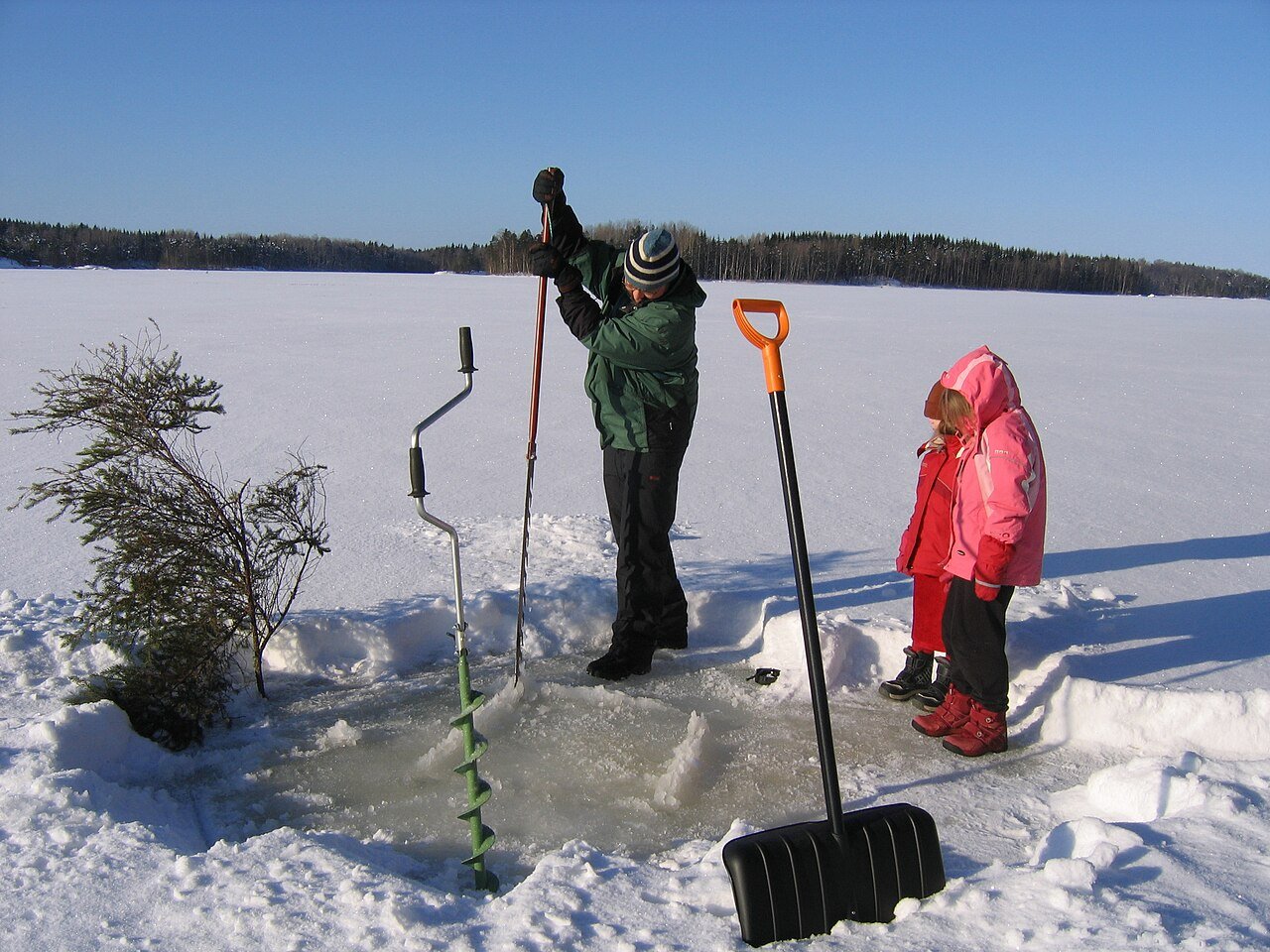 Angler drilling a hole through lake ice with an auger while a child watches